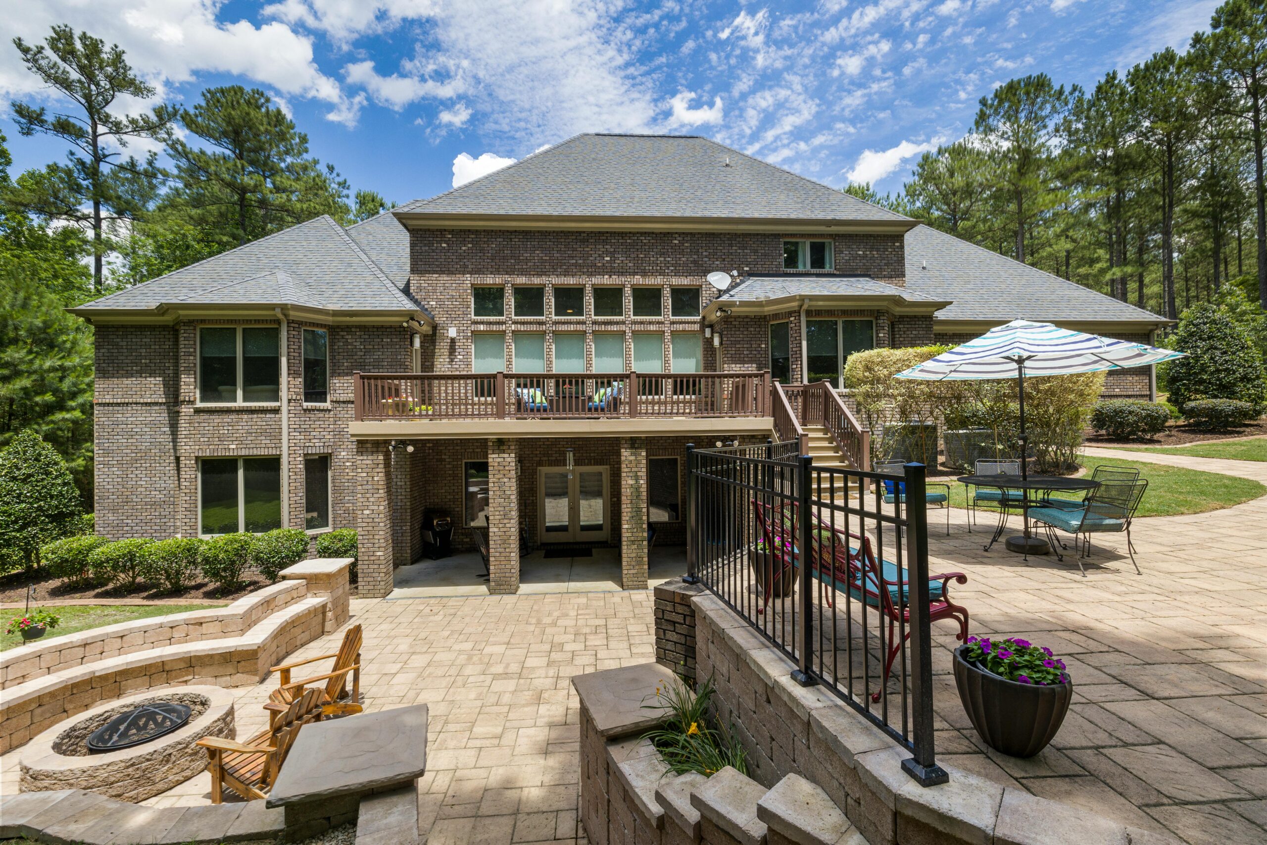 Two-story brick house with a large wooden deck, outdoor seating, and a striped umbrella on a sunny day in the yard.